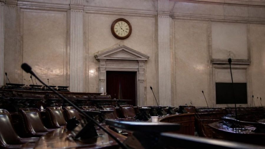 Imagen ilustrativa generada con IA: pleno legislativo vacío con micrófonos y un reloj de pared
