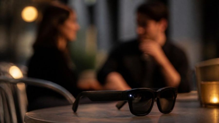 Imagen ilustrativa generada con IA: lentes inteligentes con cámara encendida sobre una mesa, con dos personas conversando al fondo